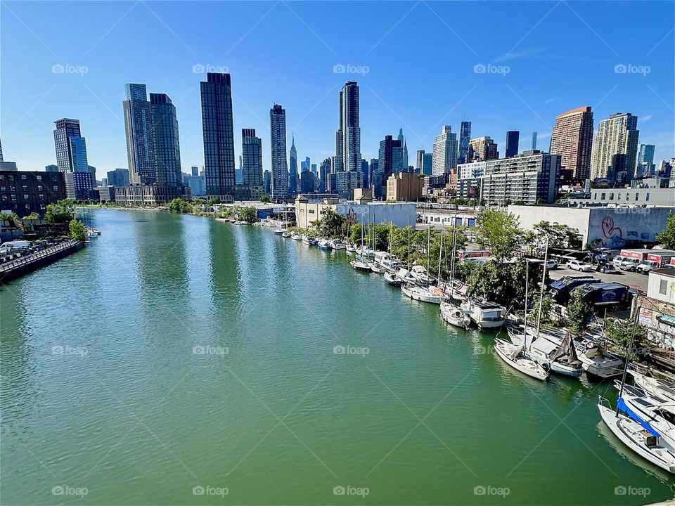 Beautiful “Newtown Creek” in turquoise with a small marina presents itself from the “Pulaski Bridge” that connects “Greenpoint”, Bklyn to LIC, Queens. In the distance we see “Manhattan” incl. the “Empire State Bldg”. 2024. Hypnotic Productions
