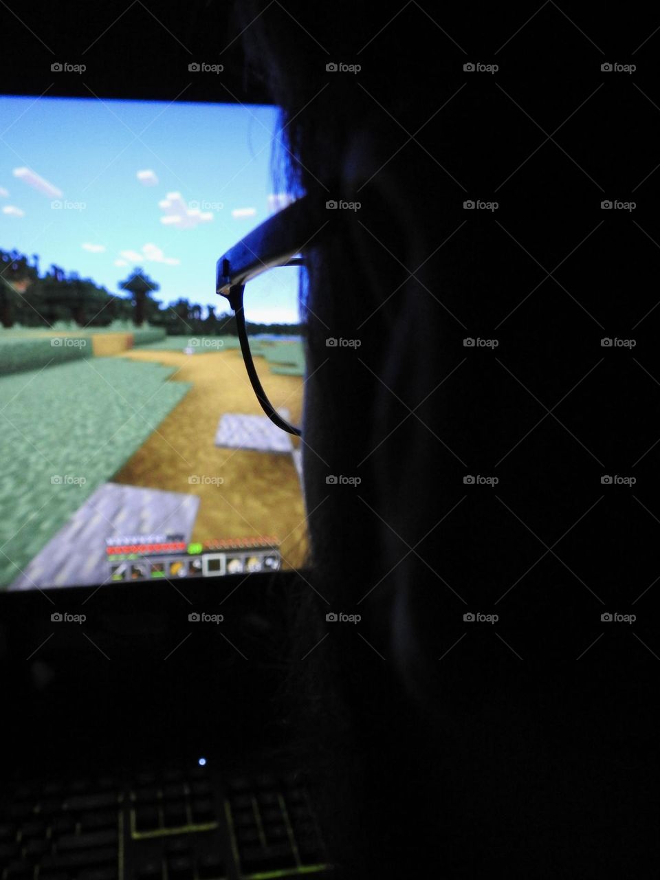 Teenager boy playing Minecraft on the gaming computer seen from his glasses close-up in a dark room.