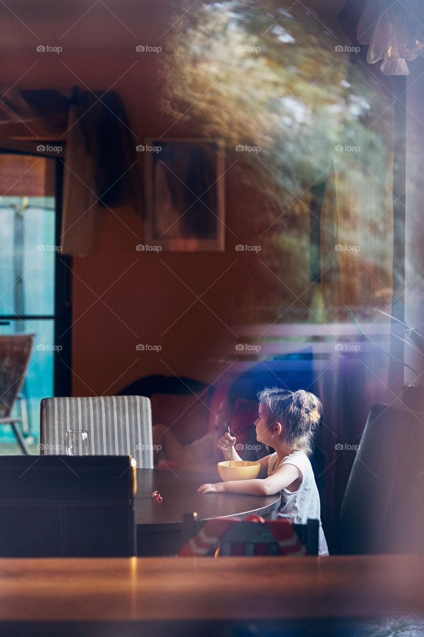 Little cute girl eating a breakfast sitting by the table. Reflections in a glass of window. Candid people, real moments, authentic situations