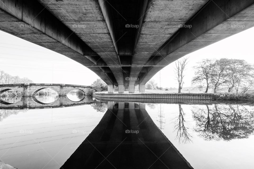 Under a modern bridge with an old, stone bridge in the background. A black and white image.