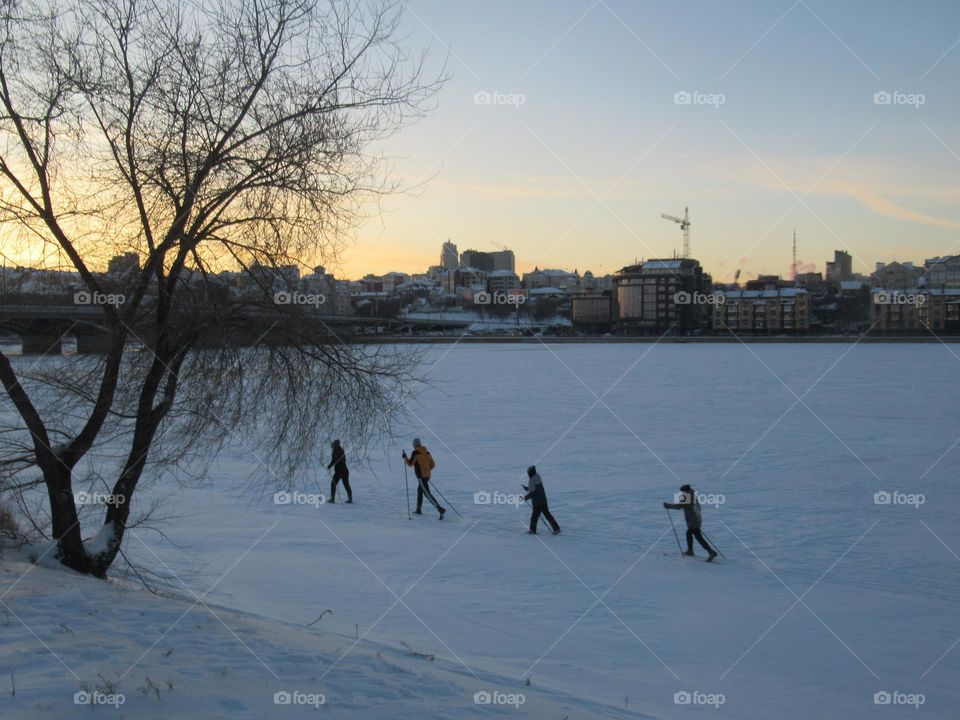 A group of skiers is walking along a frozen river, on snow and ice, winter, cold, frost, evening