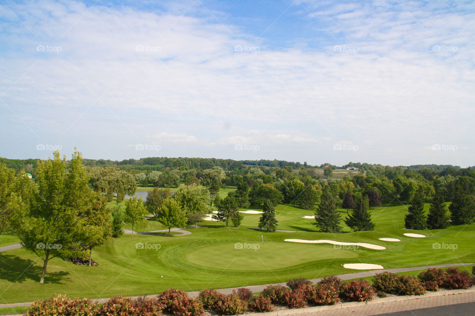 A sprawling perfect golf course with lush green grass, perfect gardens, trees and a small pond are visible from an escarpment with a blue sky dotted with white clouds