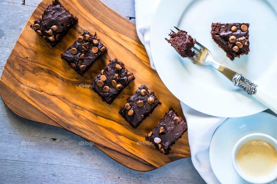 Rectangular chocolate chip brownies on a wooden serving board with a white plate and cup and saucer on the side.