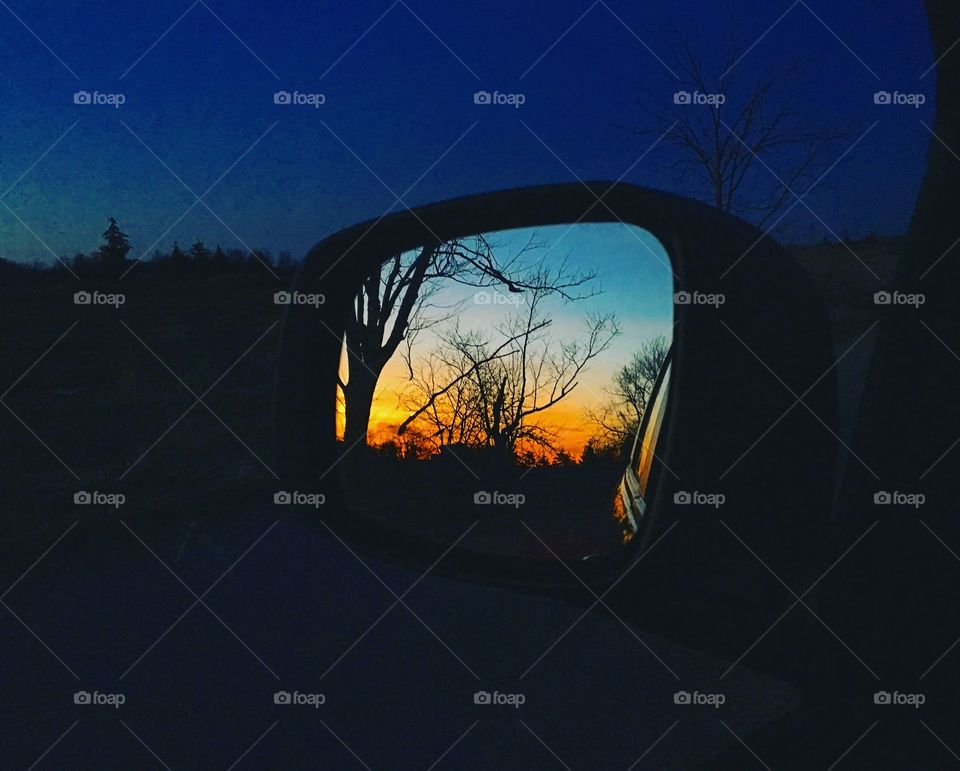A vibrant sunset with silhouettes of trees is seen in a side mirror of a car  against a deep blue sky. 