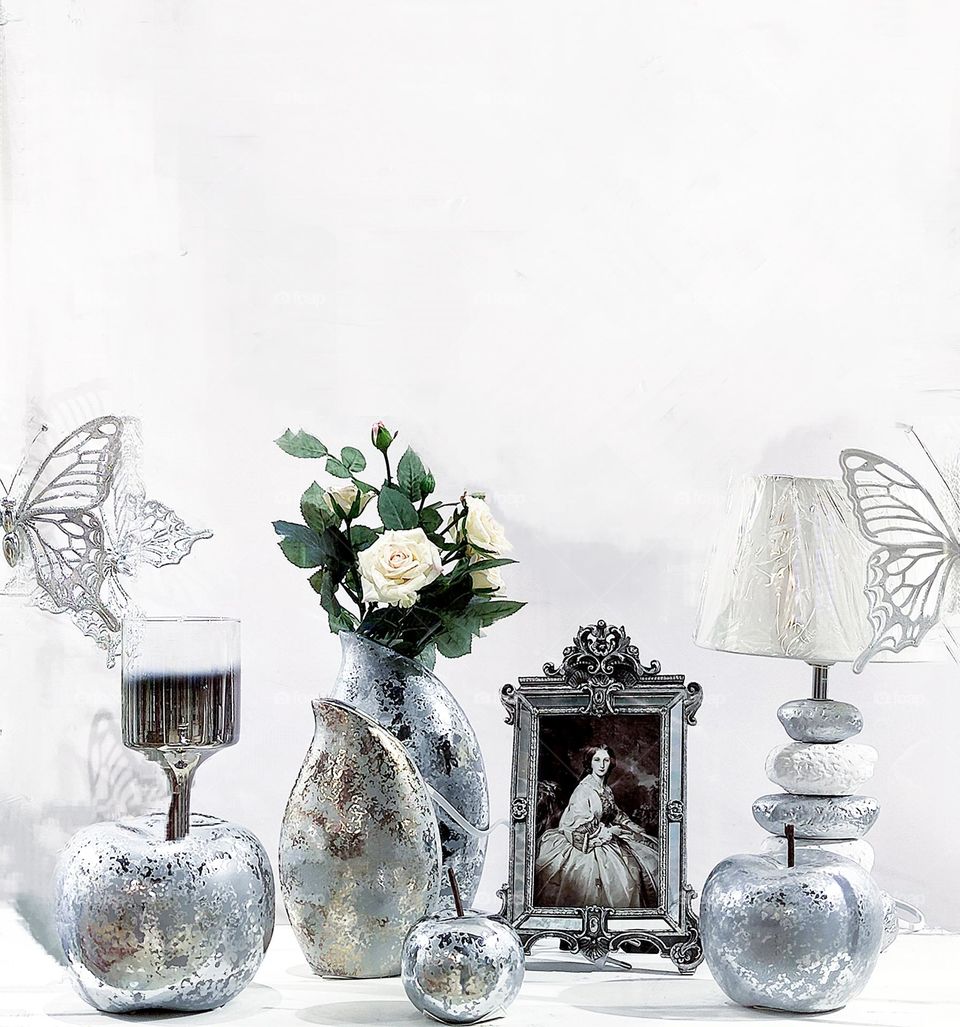 Two silvery butterflies, a glass with a silver-colored ceramic stand in the shape of an apple, a silver-colored ceramic vase with elements gold, a silver-colored ceramic apple, a frame with a portrait and a white lampshade with a ceramic stand.