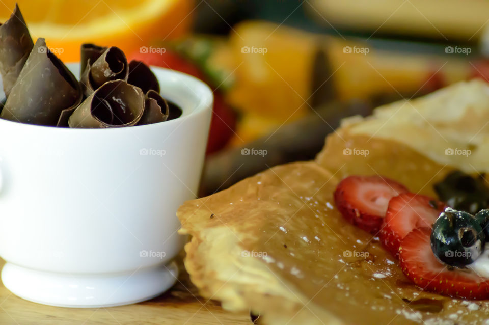 Pile of chocolate curls in porcelaine bowl next to crepe willed with fresh fruit berries and whipped cream making traditional brunch using chocolate ingredients