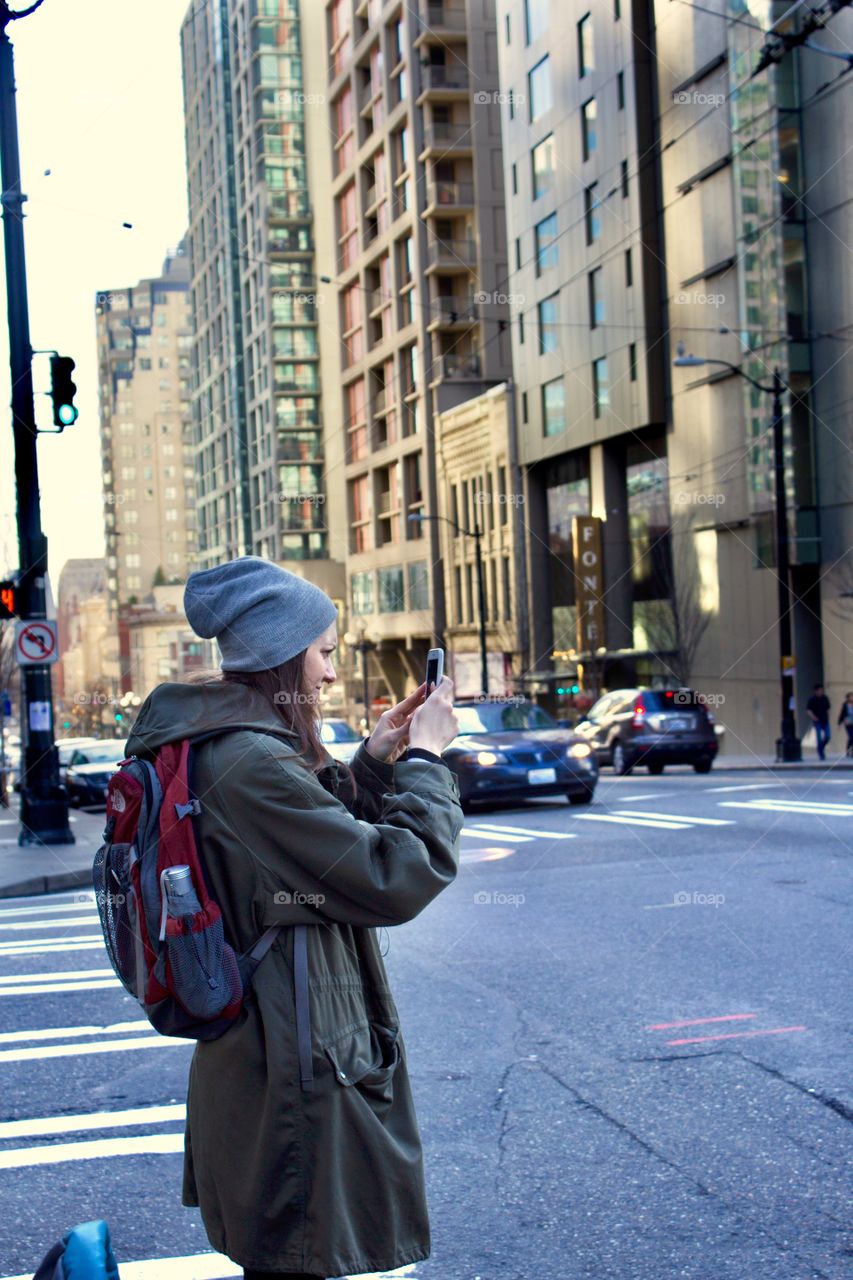 Woman in a long coat standing on the side of a busy urban street taking a photo on her cell phone