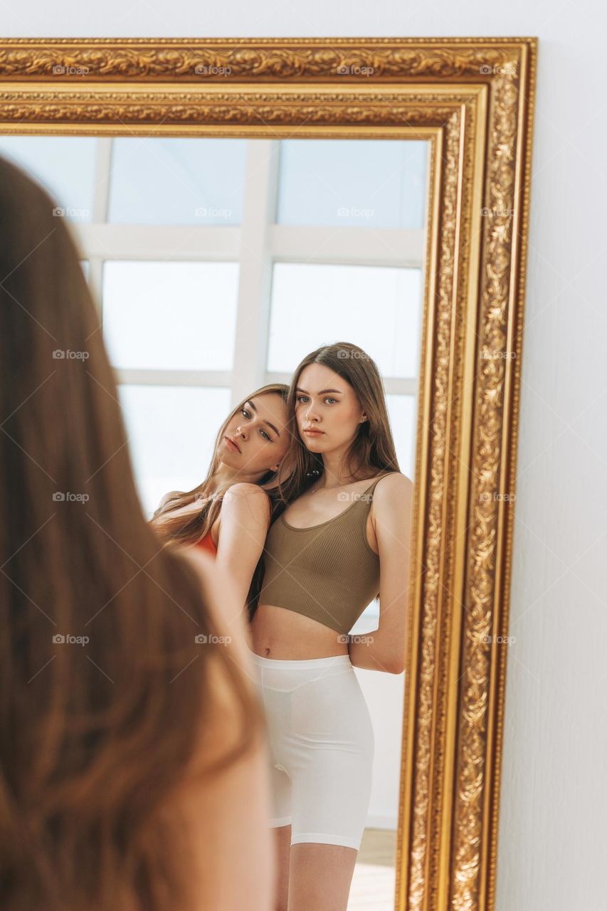 Fashion beauty models two sisters twins beautiful girls with long hair in sport wear looking to mirror in the bright studio