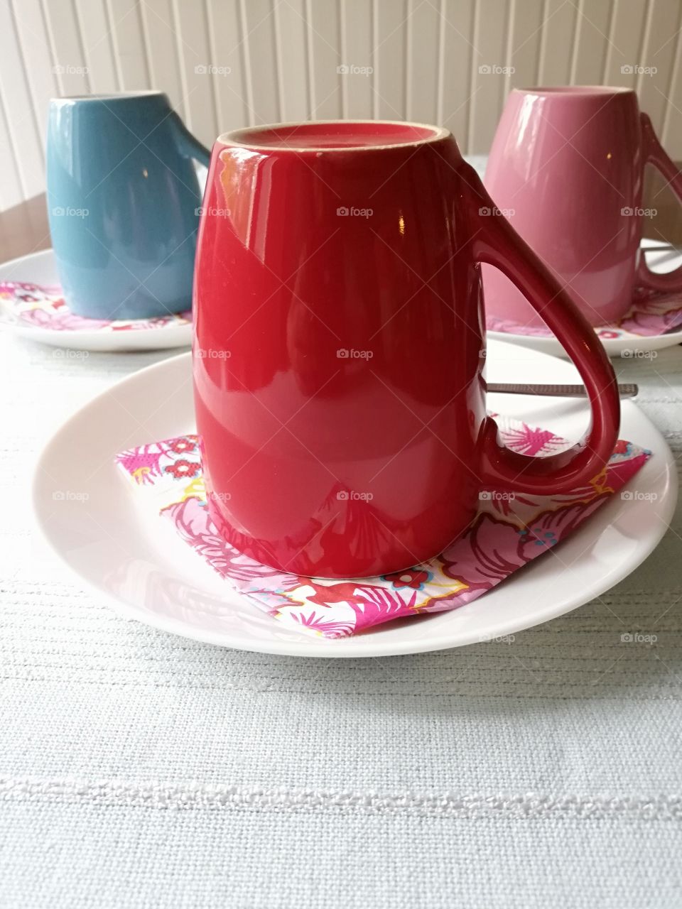 Three mugs and metallic teaspoons on the patterned napkins on the white glass plates on a light blue embossed table cloth on the brown wooden table. In the background a white painted panelled wall.