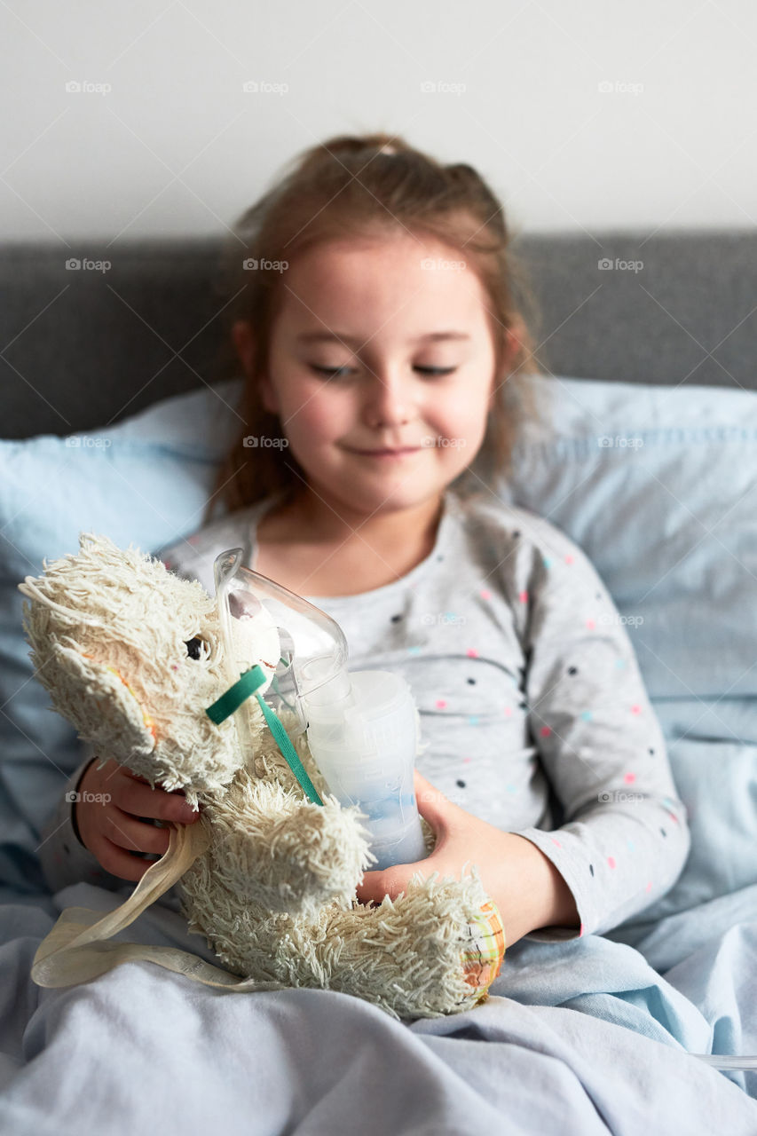 Sick child recovering in bed. Little girl playing by applying medical inhalation treatment with nebuliser to her teddy bear