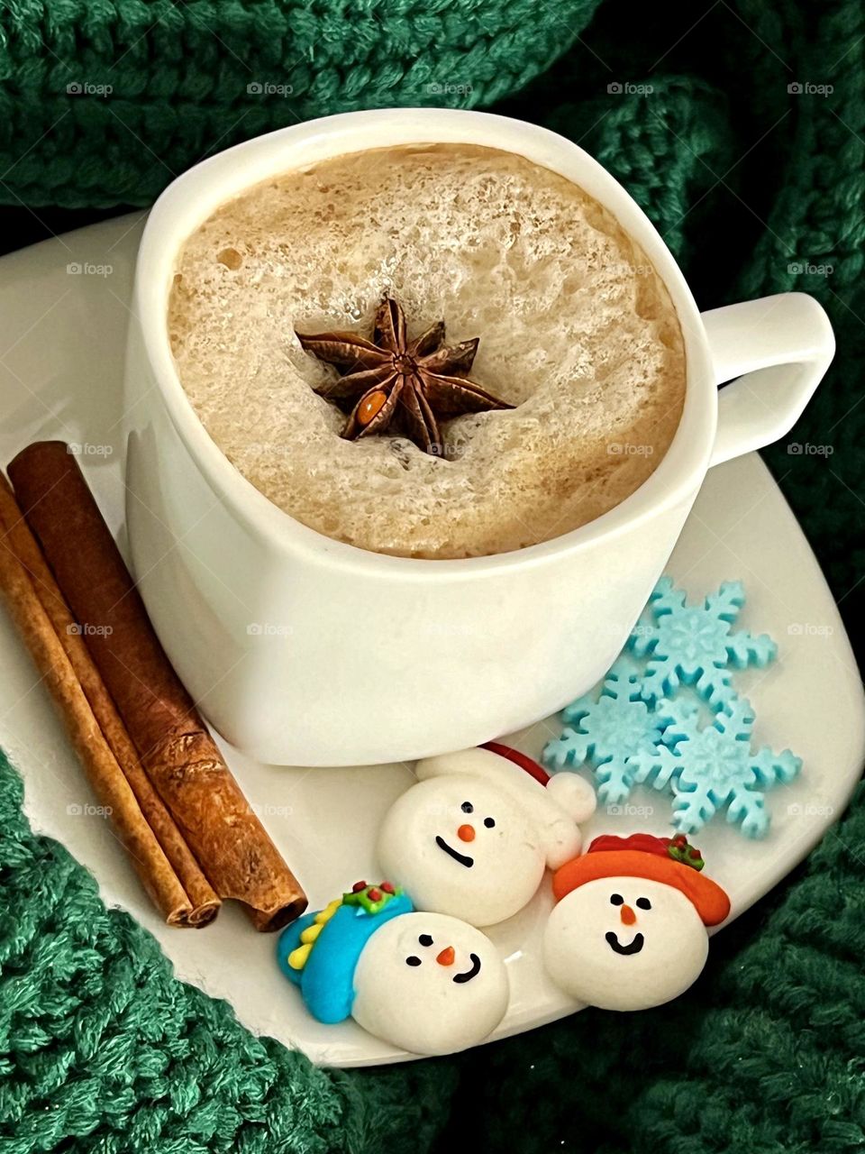 A white cup of coffee with star anise on top on the delicious foam, cinnamon on the saucer and small meringue cakes in the form of snowmen. The cup stands on a green knitted scarf.