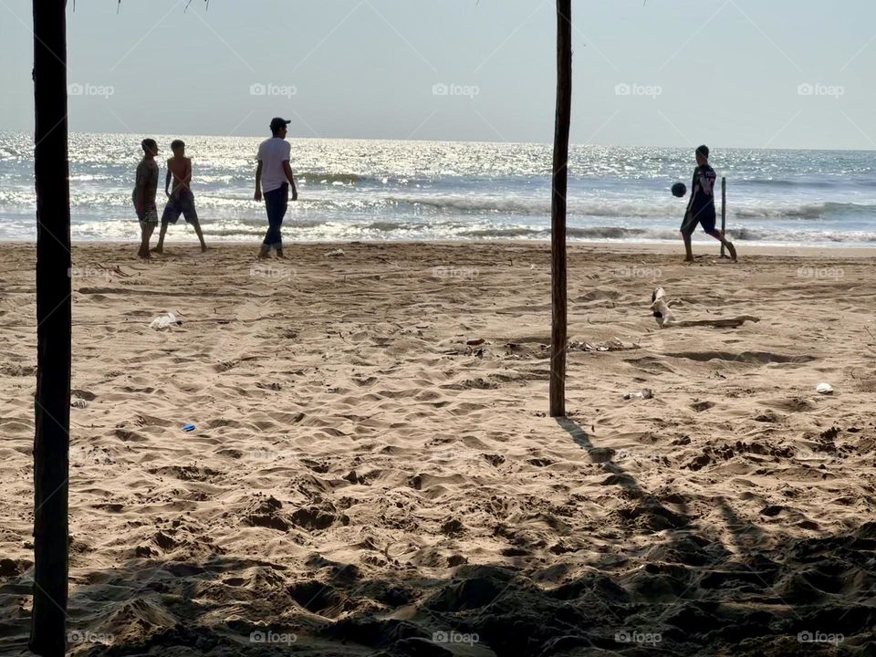 Boys playing football soccer on the beach, while the sun hits the beachfront in a sunny day.