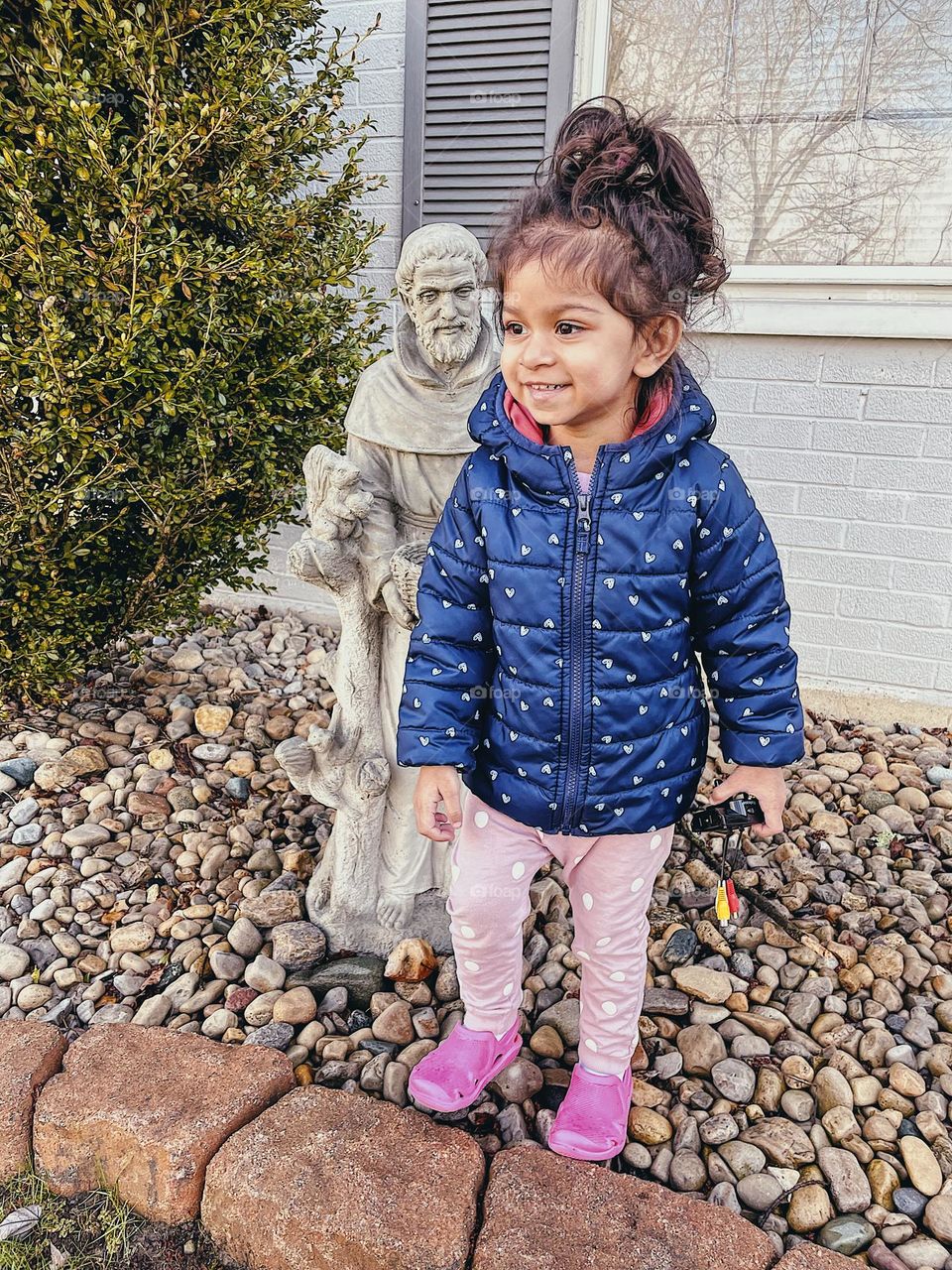 Toddler girl smiles with St. Francis statue, cement St. Francis Statue in front yard, little girl loves playing outside, interacting with statues, St. Francis  decor, religious icons, St. Francis statue looking over toddler’s shoulder 