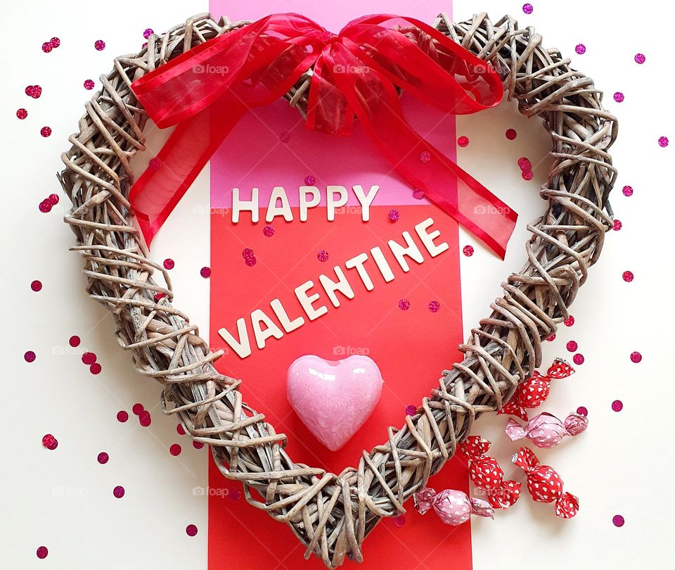 Happy valentine flatlay on cream surface composed of a heart-shaped wicker wreath with a red ribbon, a pink heart-shaped bath soap, pink and red foil coated chocolates and wooden letters placed on pink and red papers
