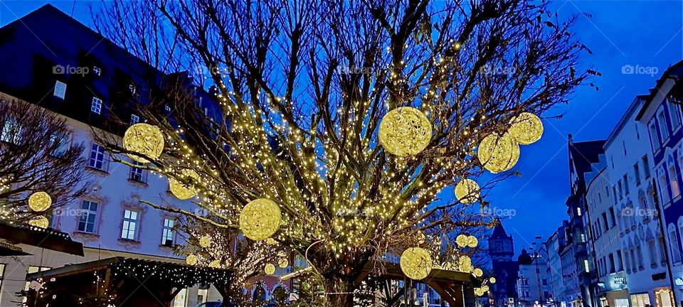 Behind the oversized illuminated Christmas tree on the “Theresienplatz”, a main square in “Straubing”, “Bavaria” sparkle lit up discs of various sizes. This is all part of the “Christkindlmarkt”, a popular Christmas fair. 2023. Hypnotic Productions