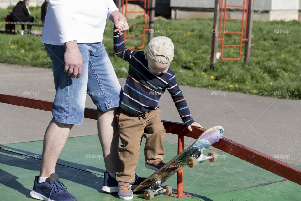 A caring and loving father patiently and calmly teaches his little son how to skateboard in an outdoor park.