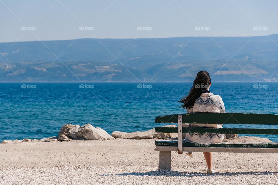 Rear view of woman sitting alone on a bench by the sea