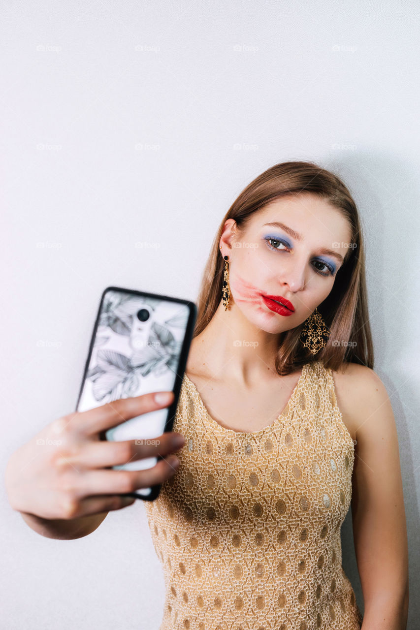 Portrait of a pretty blonde girl; her lipstick is smeared but nevertheless she looks confident and takes a selfie with her smartphone. When holds her phone in one hand and looks at its camera. She is wearing long earrings (pendants).