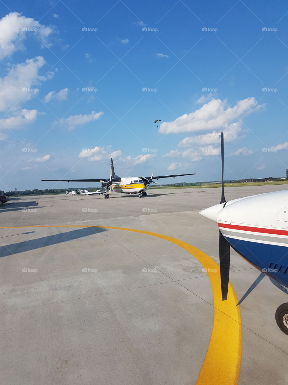 US Army Golden Knights plane on runway, prop and nose of small plane on foreground, sky diver parachuting down. Bright blue sky with fluffy clouds