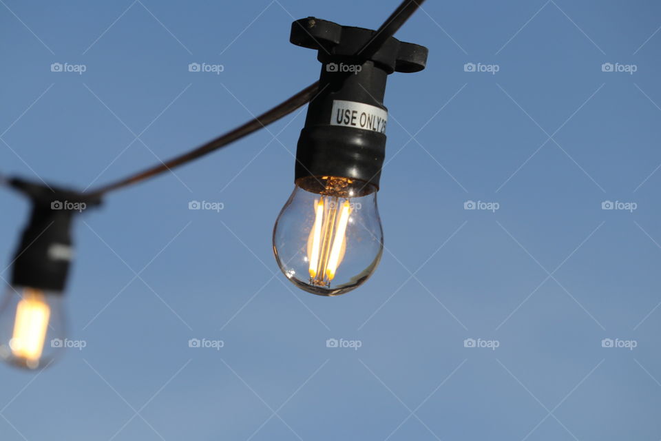 Glass electrical lightbulb globe hanging on wire illuminated in daytime against backdrop of blue sky