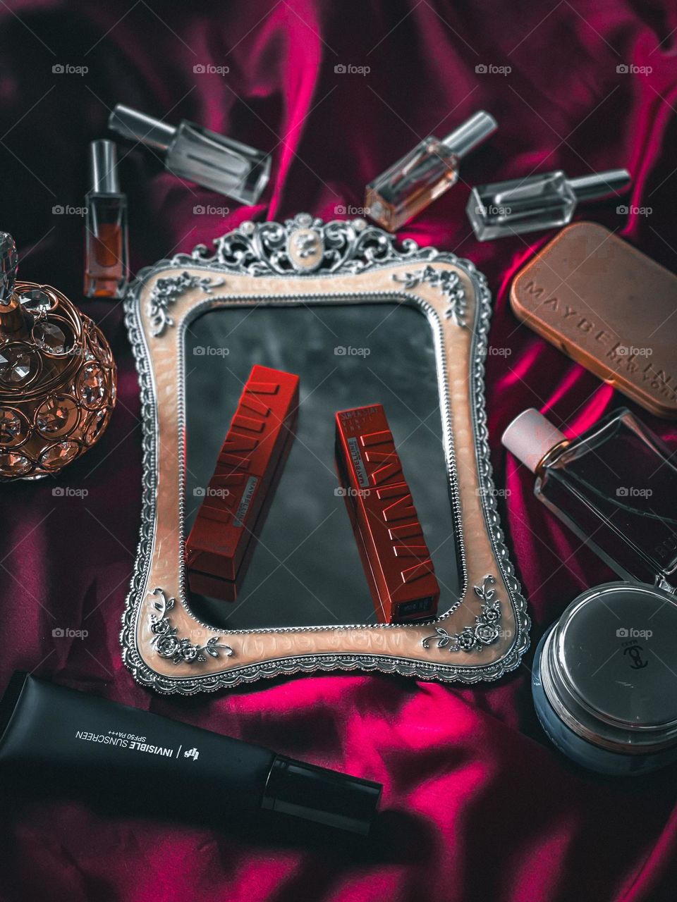 Cosmetic and skin care products on a red cloth and the center is a mirror and lipsticks on it