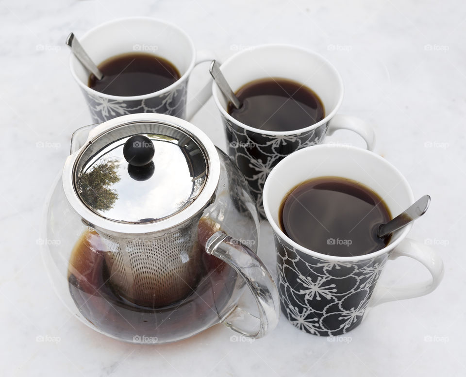 Three black and white mugs filled with strong black tea beside a round glass teapot.