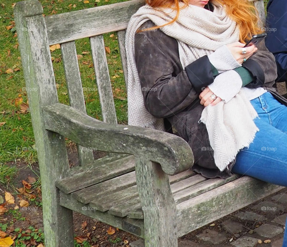 Young redhead woman sitting on wood bench on fall day and looking at her phone.