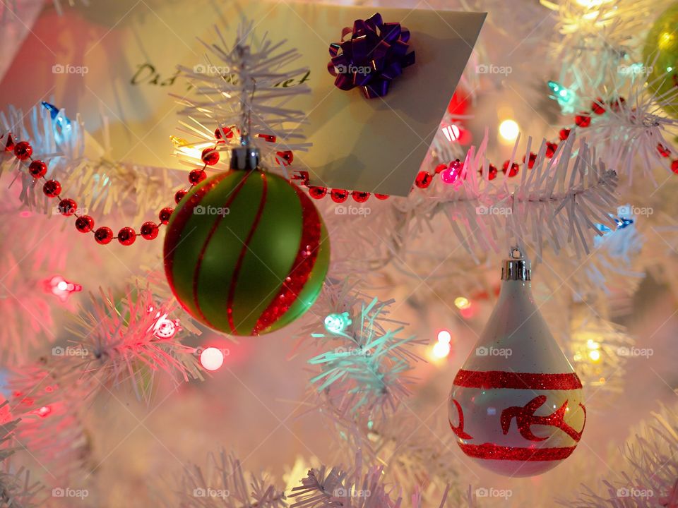 A couple of green, white, and red ornaments hanging from a white Christmas tree with soft glowing colored lights. 