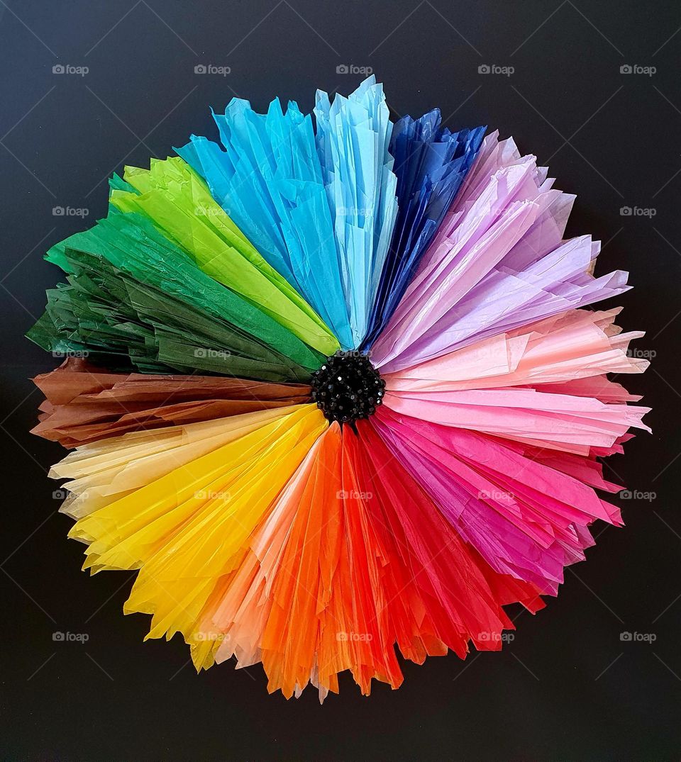 Flatlay composition of a multi-coloured flower made of silk papers and black beads placed on black paper, creating a colour explosion