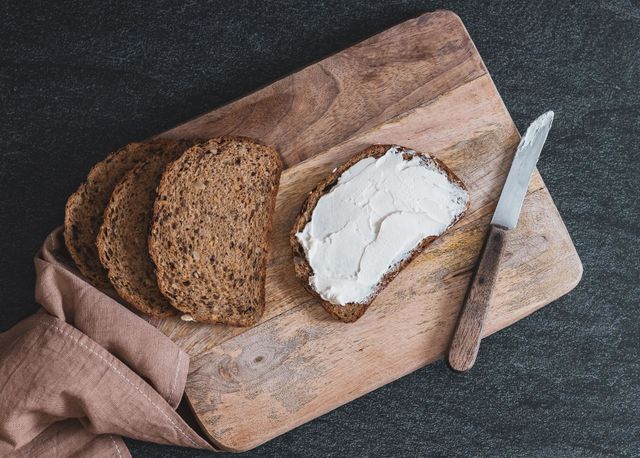 Torn pieces of bread with sunflower seeds spread with cheese mass on a wooden cutting board with a kitchen napkin and a knife lie diagonally on a chain stone background, flat lay close-up. The concept of baking bread, a healthy breakfast.
