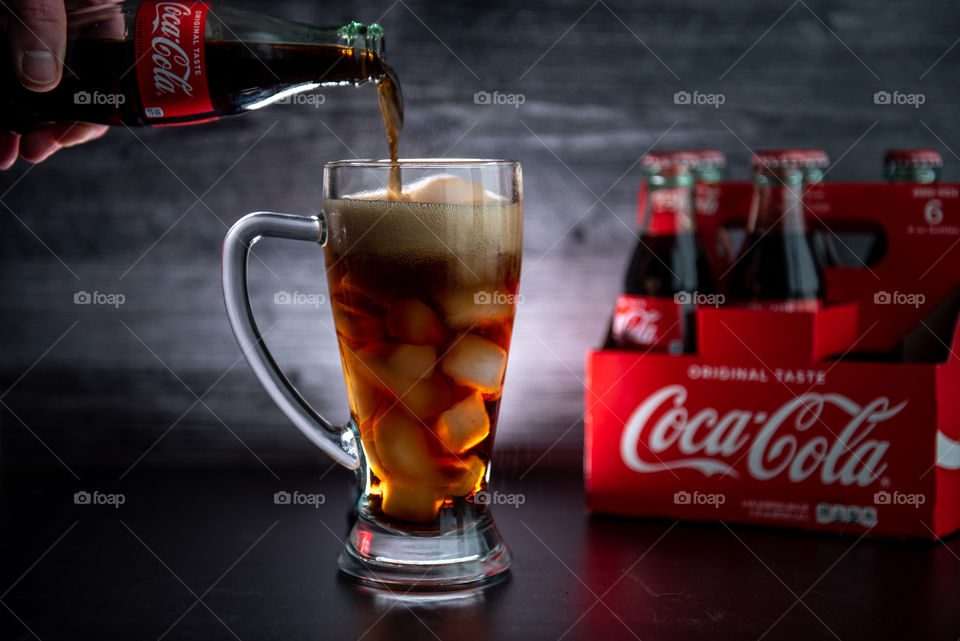 Person's hand pouring coca-cola from a bottle into a tall backlit glass with ice