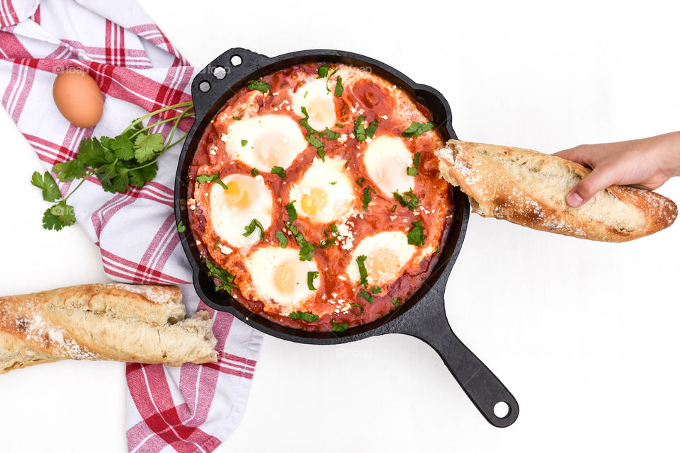 Overhead view of a person holding a baguette over a prepared shakshouka dish