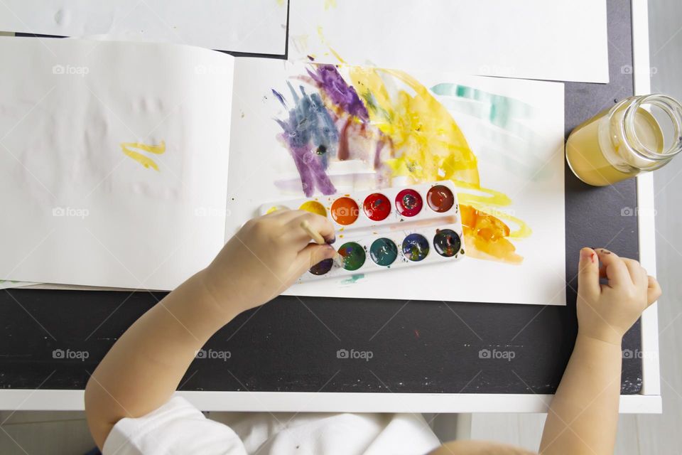 A small child draws with watercolors, multi-colored paints in an album at the table.