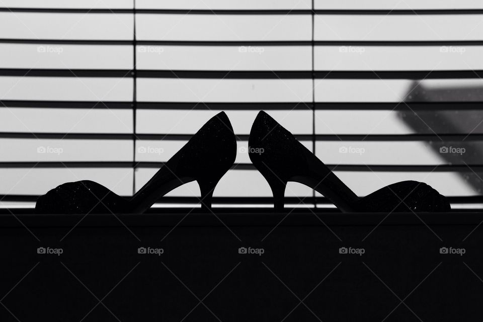 Silhouette of a bride's high heeled wedding shoes in front of a window with blinds