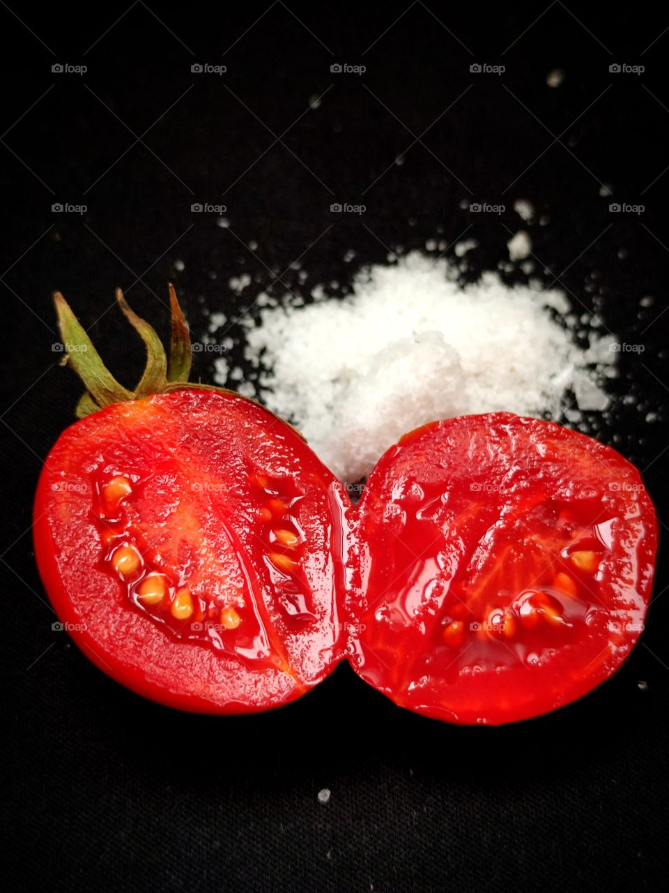 A red tomato that has been cut in half.  Nearby lies a handful of white salt.  Black background