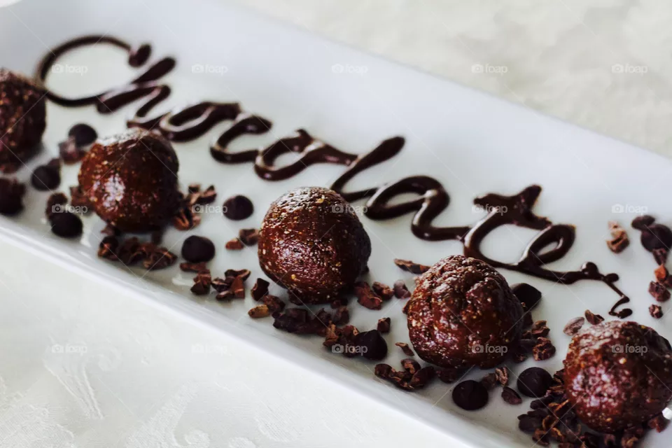 More Chocolate - Nut Butter Cocoa Bites on rectangular white dish with scattered cacao nibs and chocolate chips, and the word "Chocolate" written in chocolate drizzle