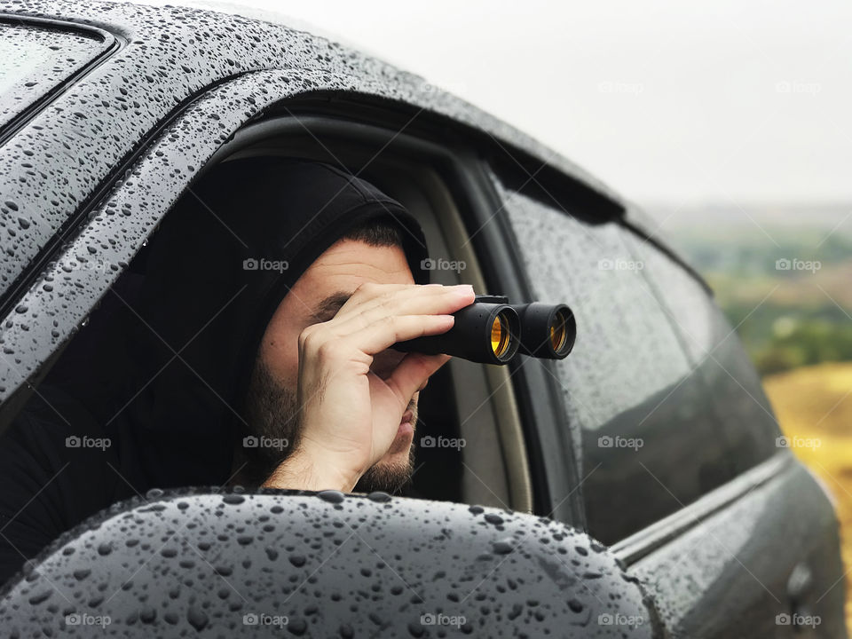 Young man spying with binoculars from the car during the rainy day 