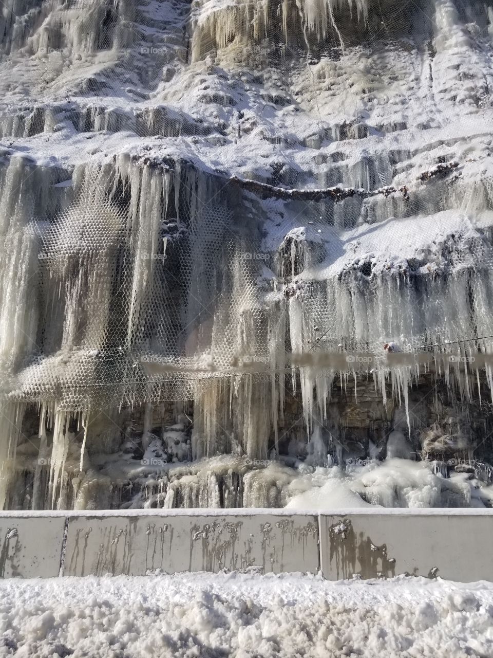 Frozen water creating the look of a frozen waterfall on a mountain-side with a chain link fence to prevent the dangers of fallen rocks.