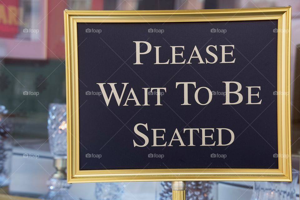 A gold color floor standing sign outside a restaurant reads “Please Wait To be seated”