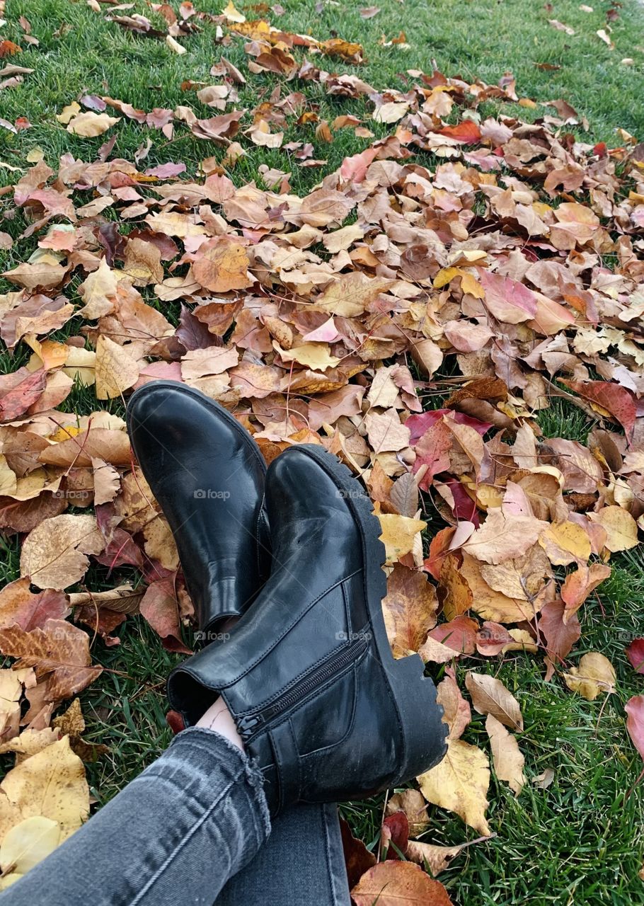 Woman wearing a pair of black boots sitting on the green grass and autumn leaves 
