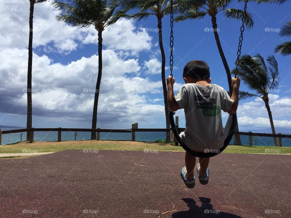A toddler sits on a swing against a beautiful backdrop of palm trees and blue sky.
