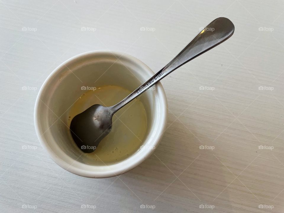 An empty ice cream cup with a silver spoon sticking out of it. The cup is on top of a white table. A shadow looks like it’s gripping the cup.