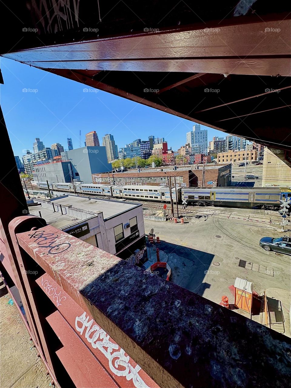This is the view of the “Long Island Railroad”, the silver train passing us by from the middle platform of red metal staircase of the “Pulaski Bridge” at “Newtown Creek” in LIC, Queens. 2024. Hypnotic Productions