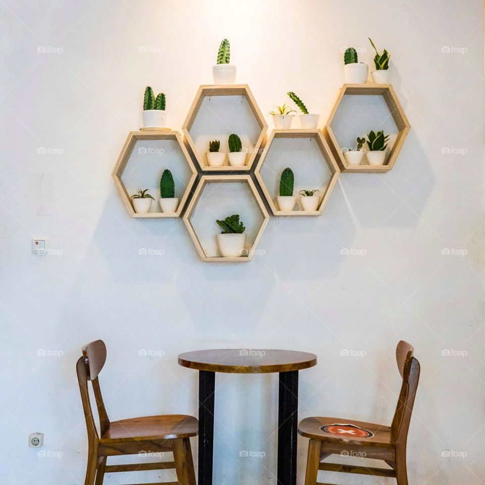 Two chairs and a table next to a wall decorated with decorative plant shelves. Chair. Table. Circle. Wood. Rack. Hexagon. Plant. Decoration. ornamental. Interiors. Café. Seat. Wall. Photography. Scene