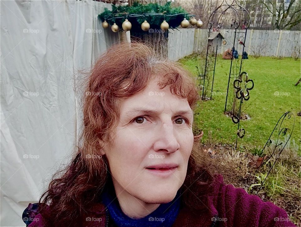 Christmas decorations are tied to a basket with potted plants and cast iron artwork beautifies the garden of a house in “Bavaria”, a flower, an arch with a birdhouse hung from it. The lovely lady is the proprietor. 2023. Hypnotic Productions