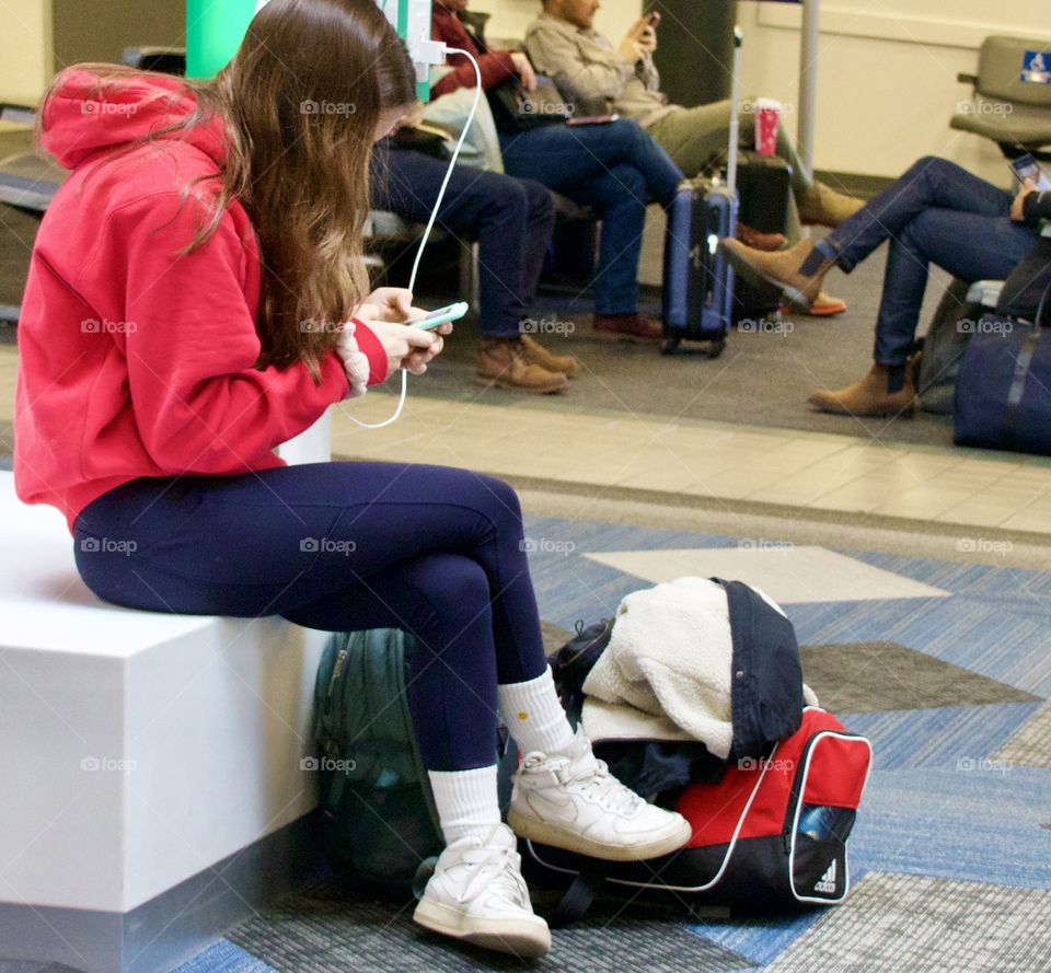 Girl wearing red and blue sitting at the airport charging her cell phone while texting