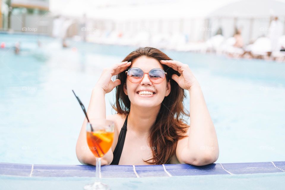 Stylish happy young woman plus size body positive in black swimsuit and sunglasses enjoying her life with glass of cocktail in the hotel pool, summer vacations