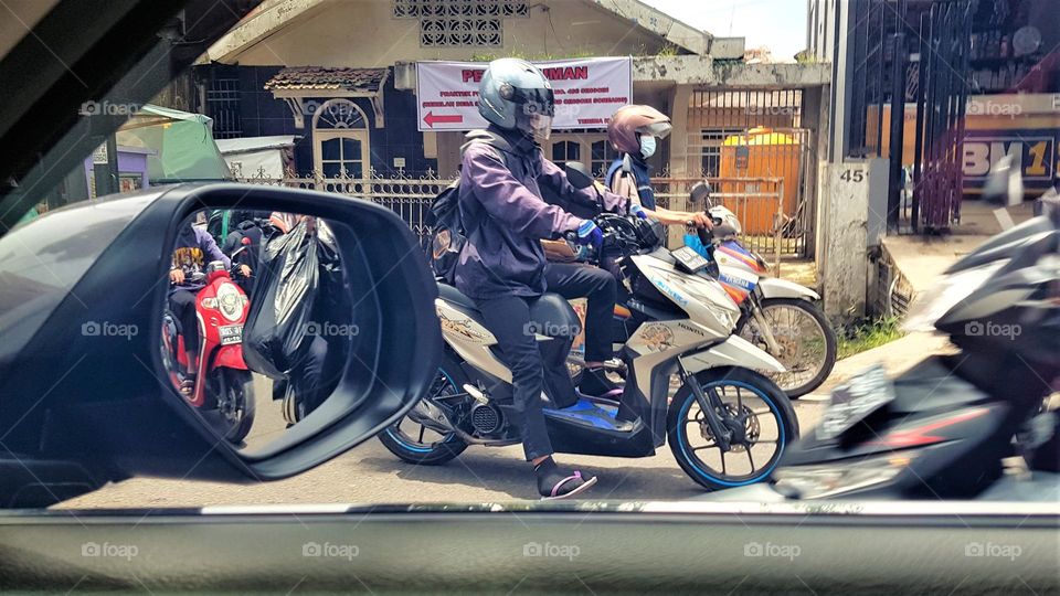 behind the car cabin, view main road, dominated by motorcycle dirver, motorbike driver wearing helmets, motorbikes are visible in rearview mirror, the road is quite busy,
