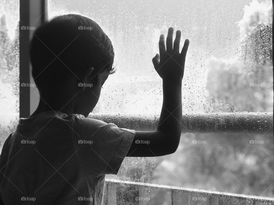 A black and white picture of a sad boy standing near window on heavy rainy day and watching outside tragedy 