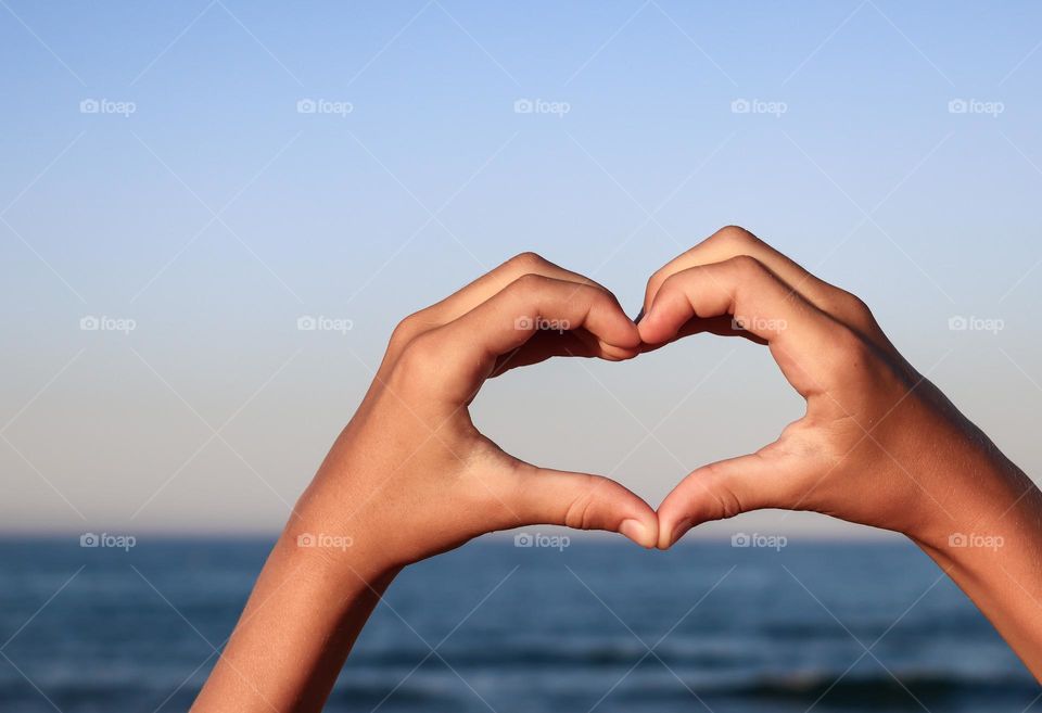 A beautiful view of children's hands forming a heart with their fingers against the backdrop of the sea on a summer sunny day, side view close-up.